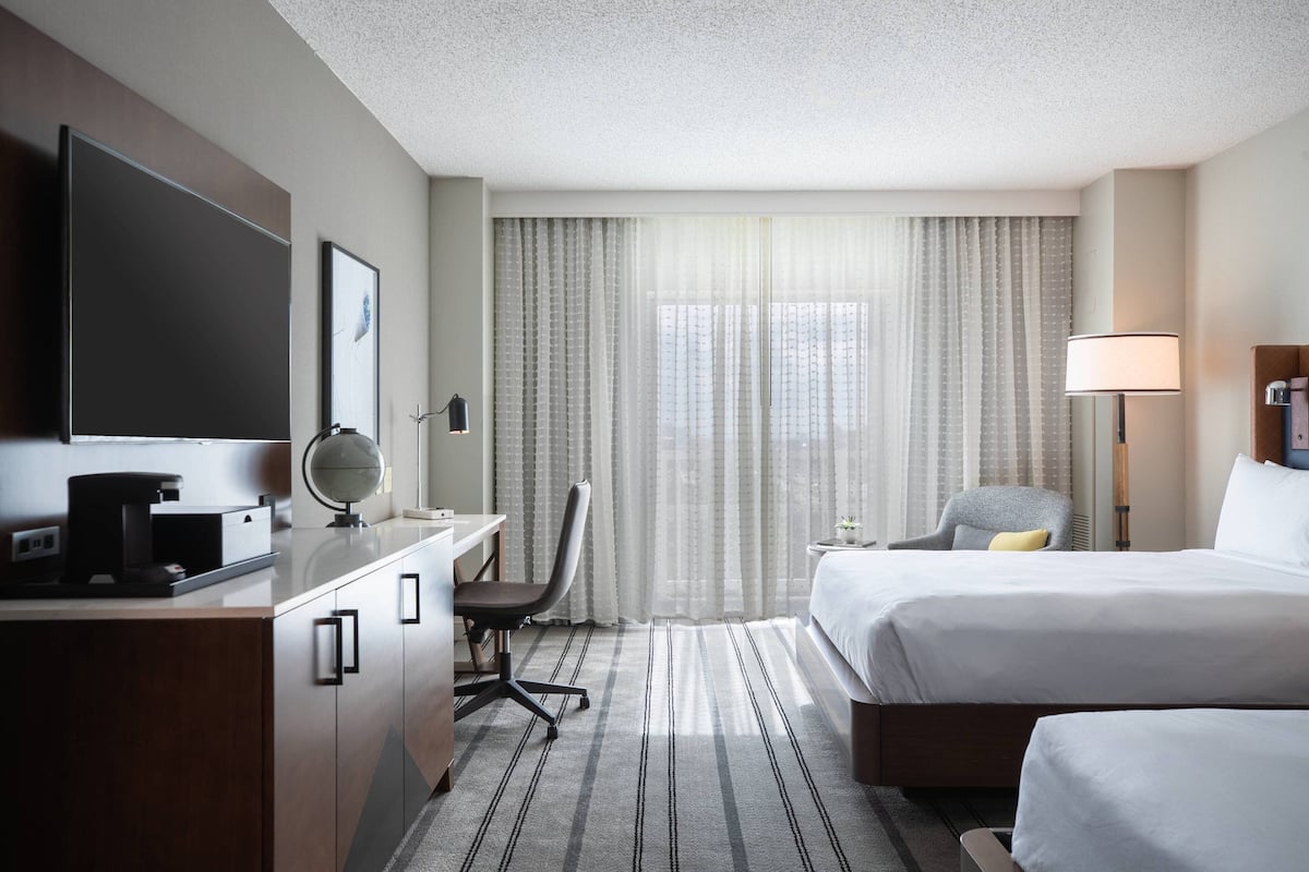 A hotel room with a bed, a desk with a computer, a chair, a TV, and a window with curtains.