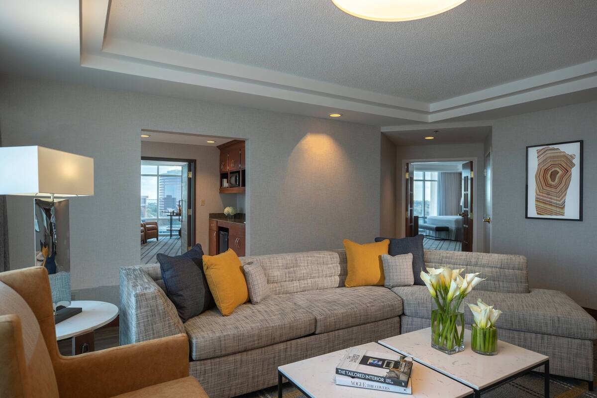 A modern living room with a sectional sofa, a coffee table, and a lamp. There is a bookshelf and a framed artwork on the wall.