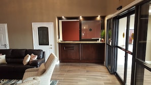 Un salon avec un canapé et un fauteuil à motifs, un coin cuisine avec un meuble rouge, et une porte-fenêtre coulissante donnant sur l’extérieur.