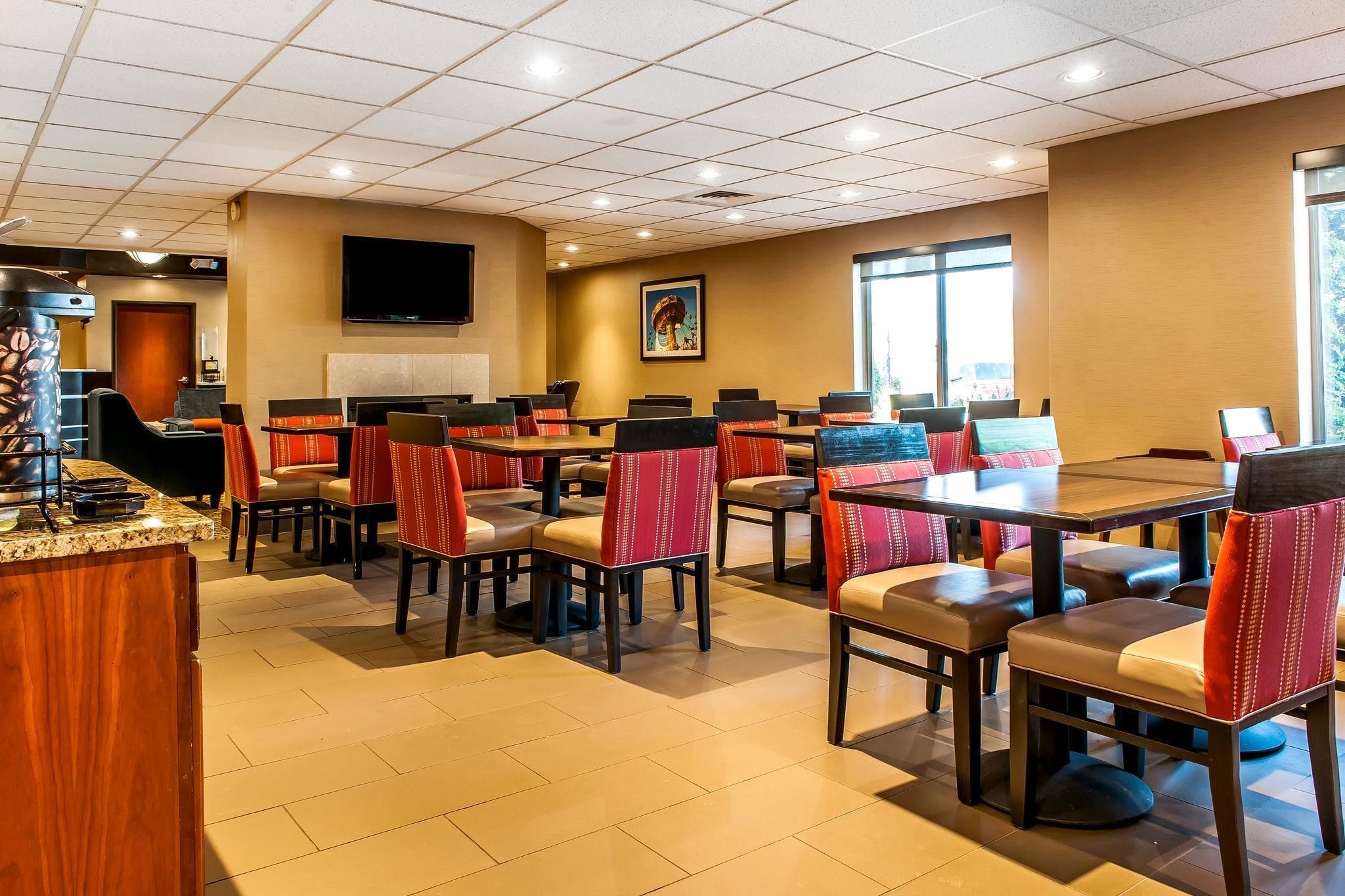 A hotel dining area with multiple tables and chairs, a television mounted on the wall, and a large window providing natural light.