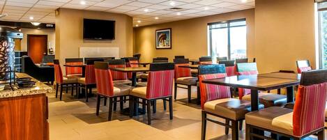 A hotel dining area with multiple tables and chairs, a television mounted on the wall, and a large window providing natural light.