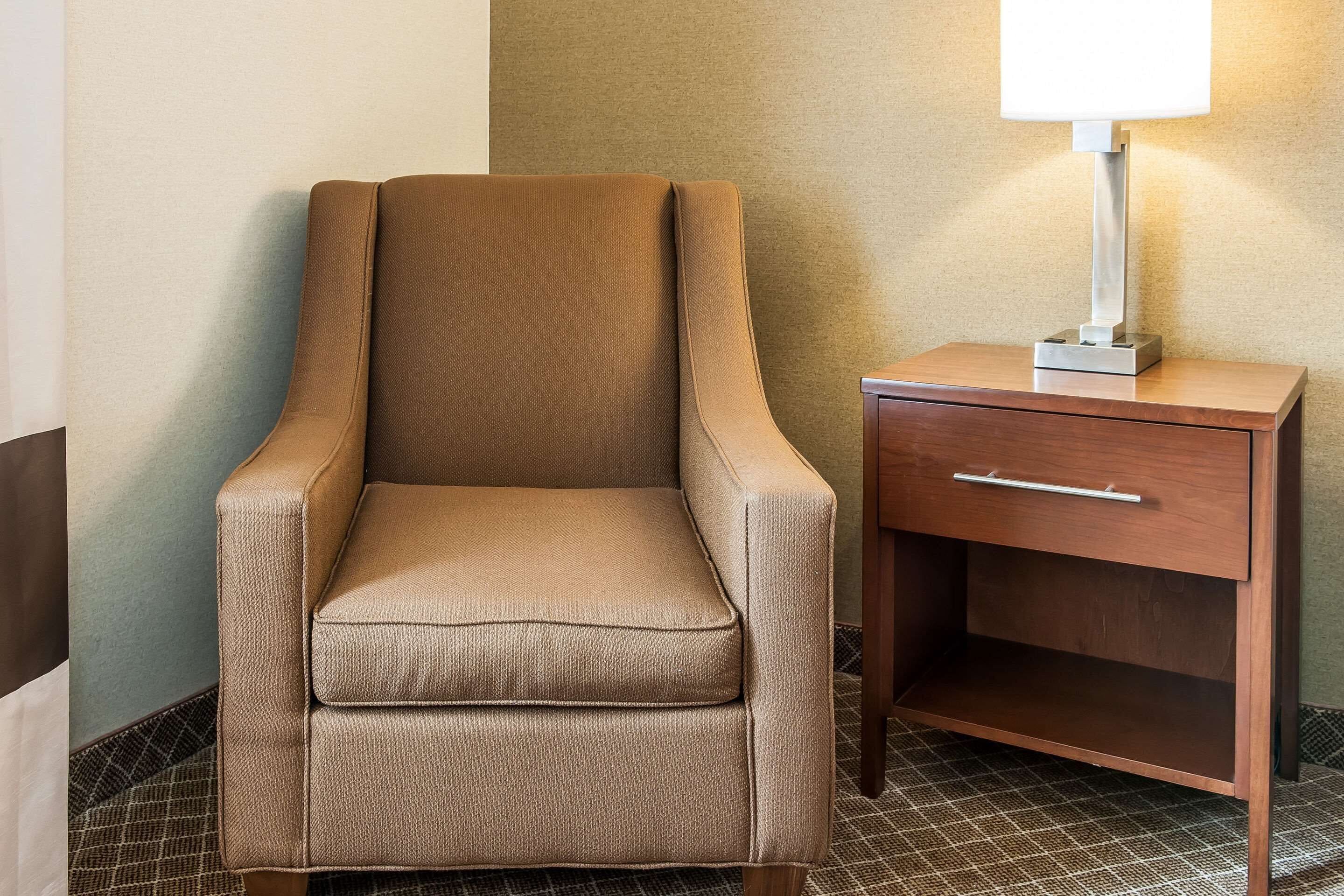A tan armchair with a wooden nightstand beside it, featuring a drawer and a lamp.