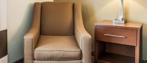 A tan armchair with a wooden nightstand beside it, featuring a drawer and a lamp.