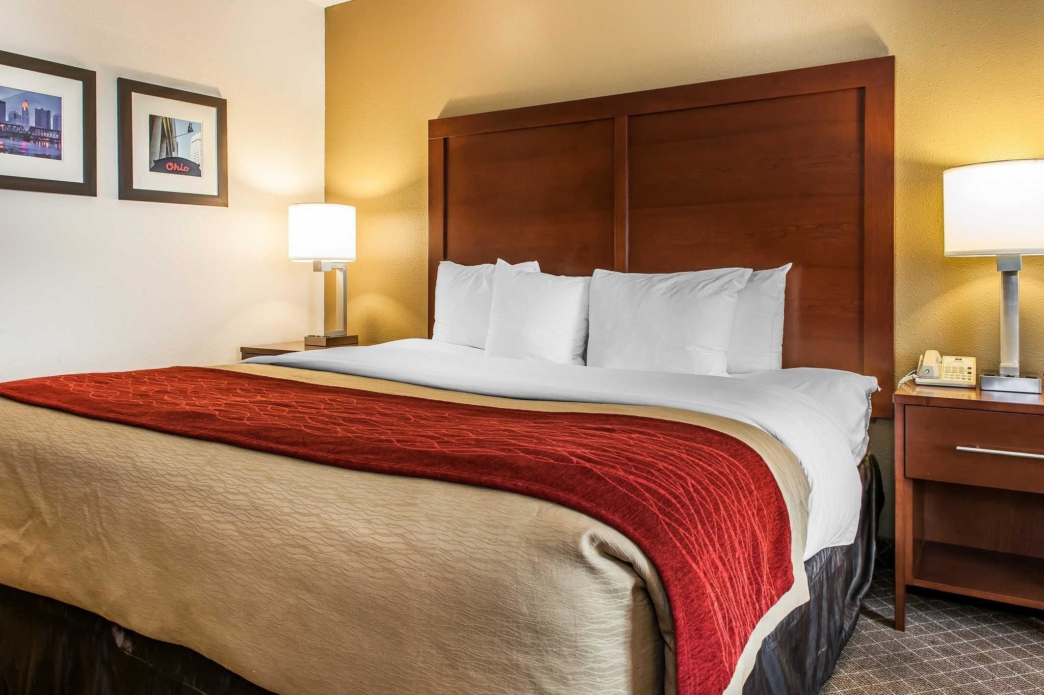 A hotel room with a large bed, a wooden headboard, two bedside lamps, and a red throw blanket.
