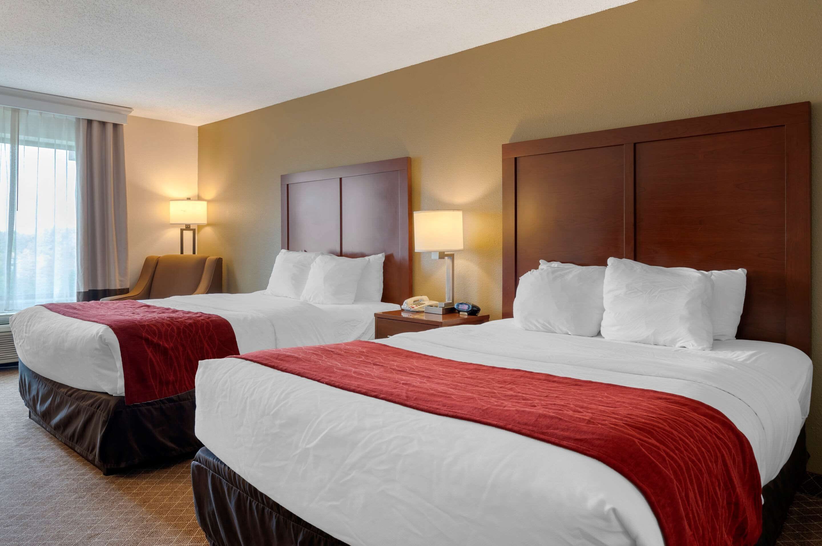 Habitación de hotel con dos camas, cada una con ropa de cama blanca y mantas rojas. Dos mesitas de noche con lámparas, una silla y una ventana con cortinas.
