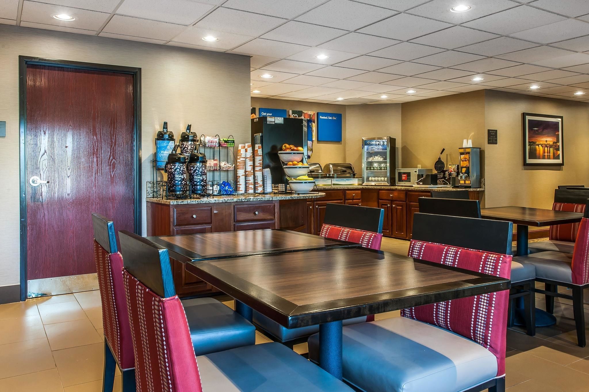 A hotel dining area with a large wooden table, blue and red cushioned chairs, a buffet counter, and a coffee machine.