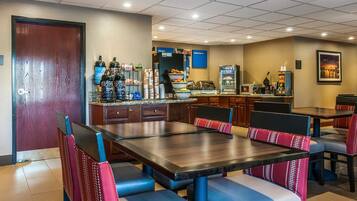 A hotel dining area with a large wooden table, blue and red cushioned chairs, a buffet counter, and a coffee machine.
