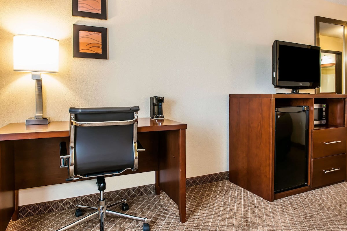 A hotel room with a wooden desk, a black office chair, a lamp, a television, a refrigerator, and a microwave.
