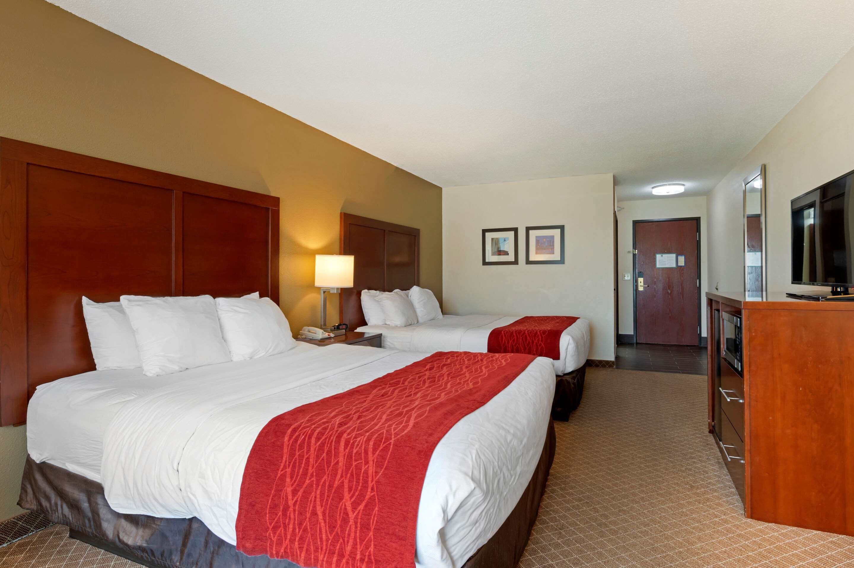 Quarto de hotel com duas camas, uma escrivaninha, uma lâmpada, uma televisão e uma porta que dá para outro cômodo.