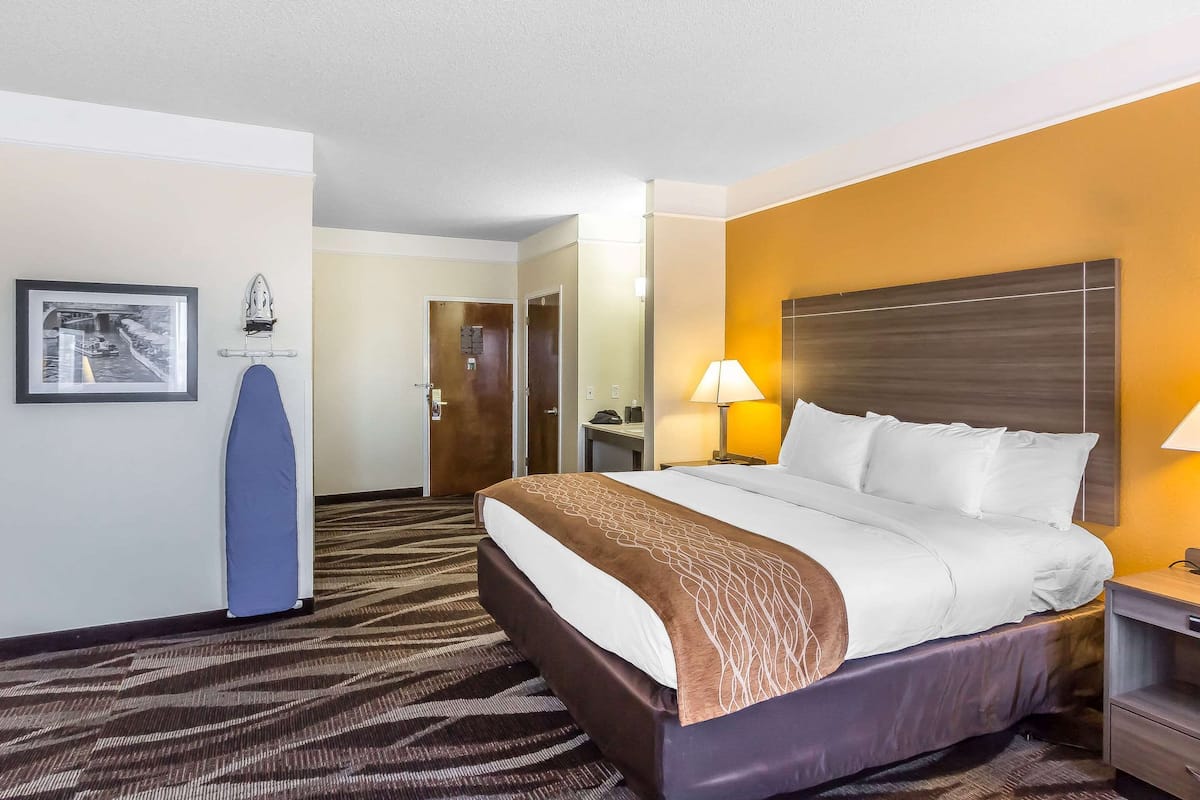 Een hotelkamer met een groot bed, een nachtkastje met een lamp, een inlijsting foto aan de muur en een deur die naar een andere kamer leidt.