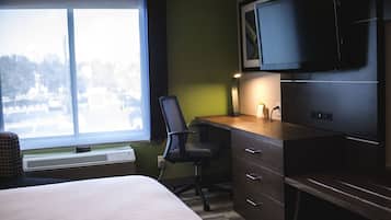 A hotel room with a bed, a desk with a chair, a television, and a window with a view of trees.