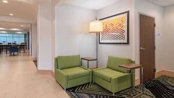 Un vestíbulo de hotel con una recepción, dos sillones verdes, una alfombra estampada y una lámpara de pared.