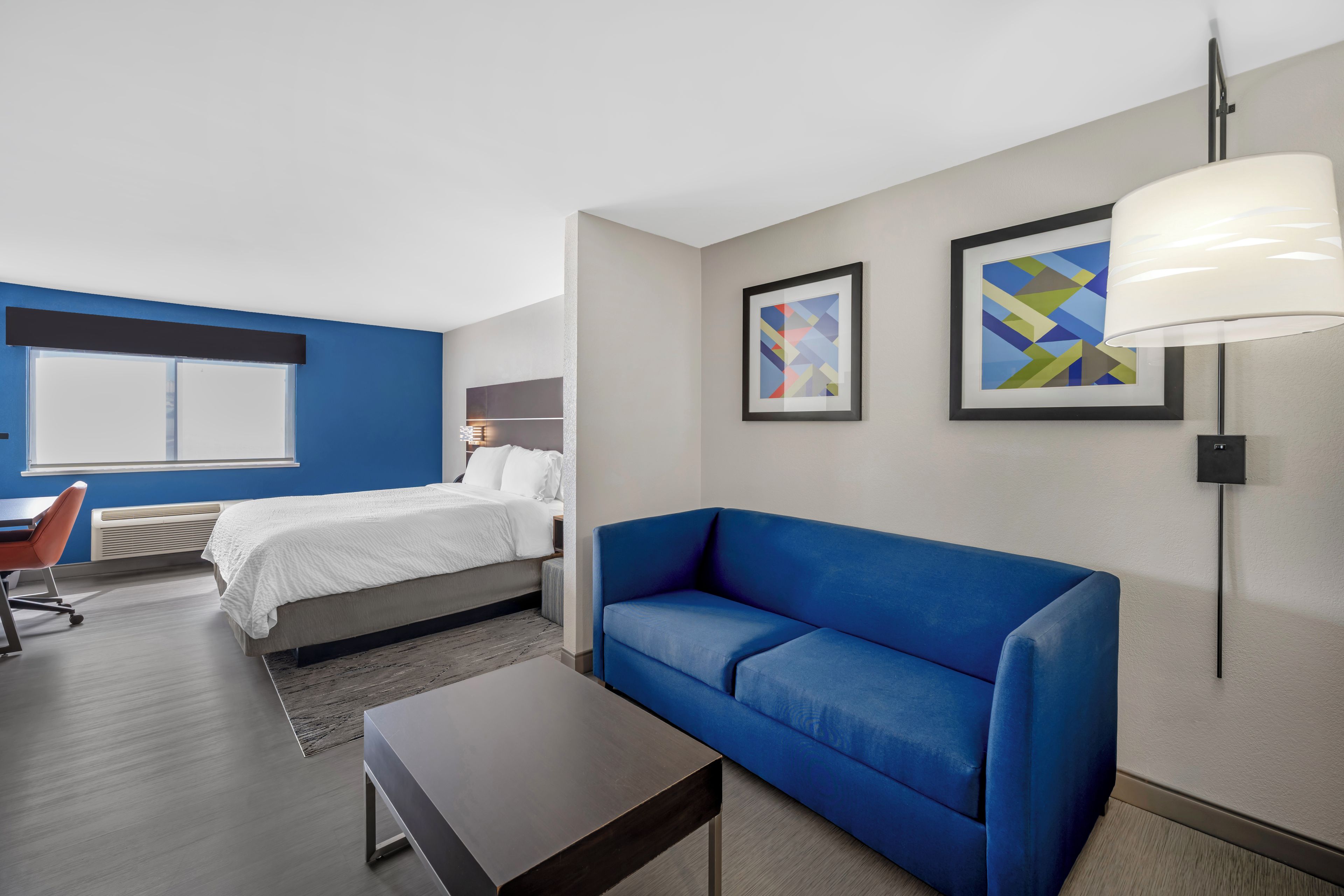 A modern hotel room with a bed, a blue sofa, a desk, and a chair. There are two framed artworks on the wall.