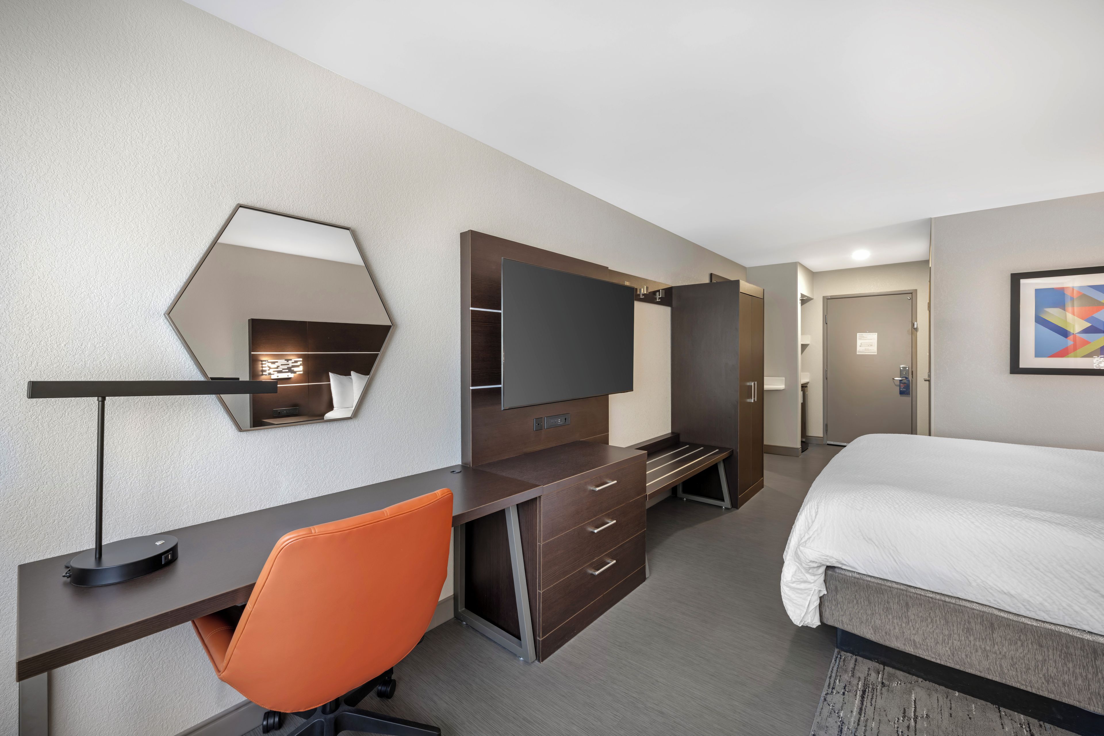 A hotel room with a bed, a desk with a chair, a television, a wardrobe, and a mirror.