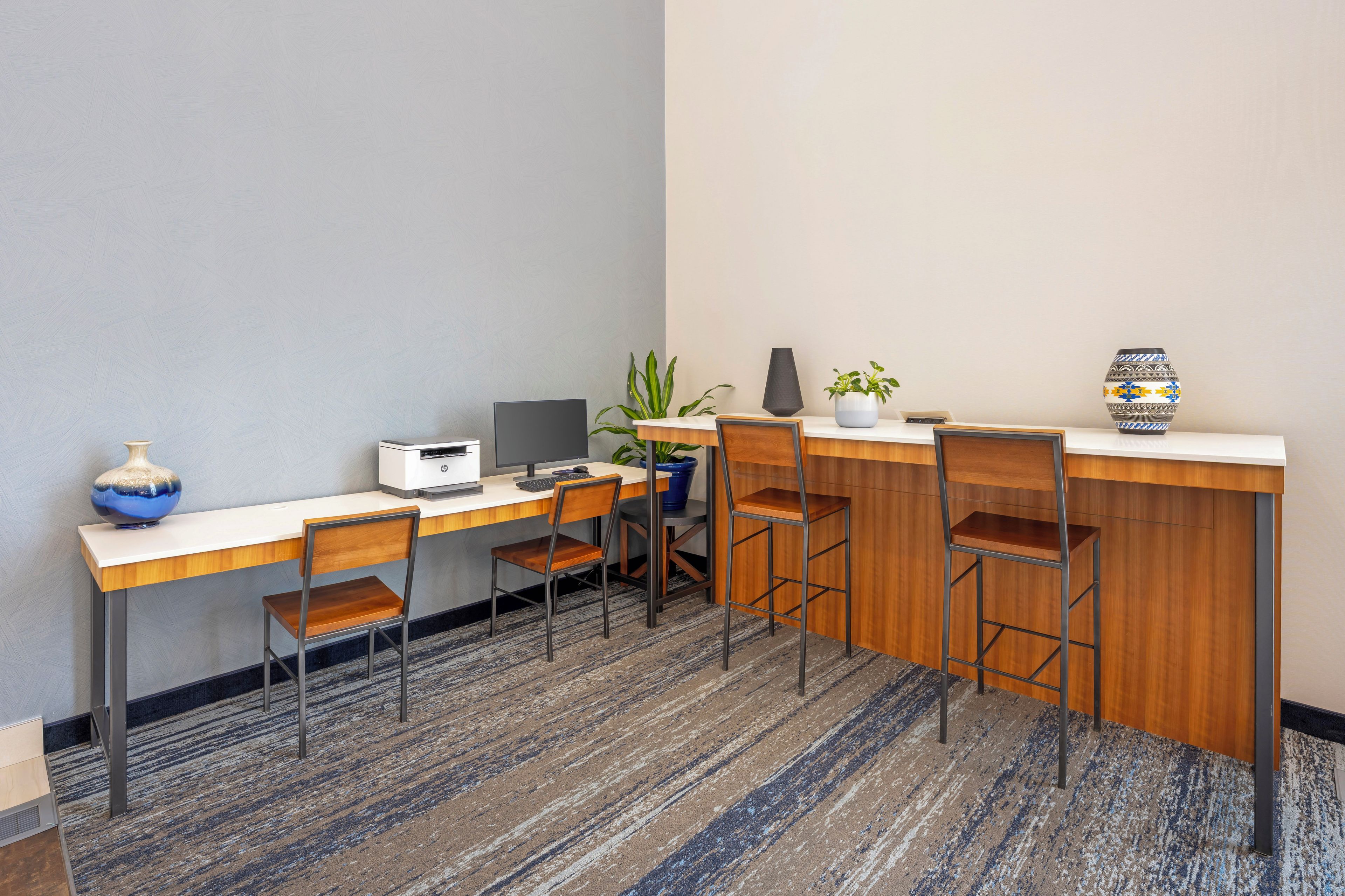 A hotel room with a desk, computer, chairs, a potted plant, and a decorative vase.