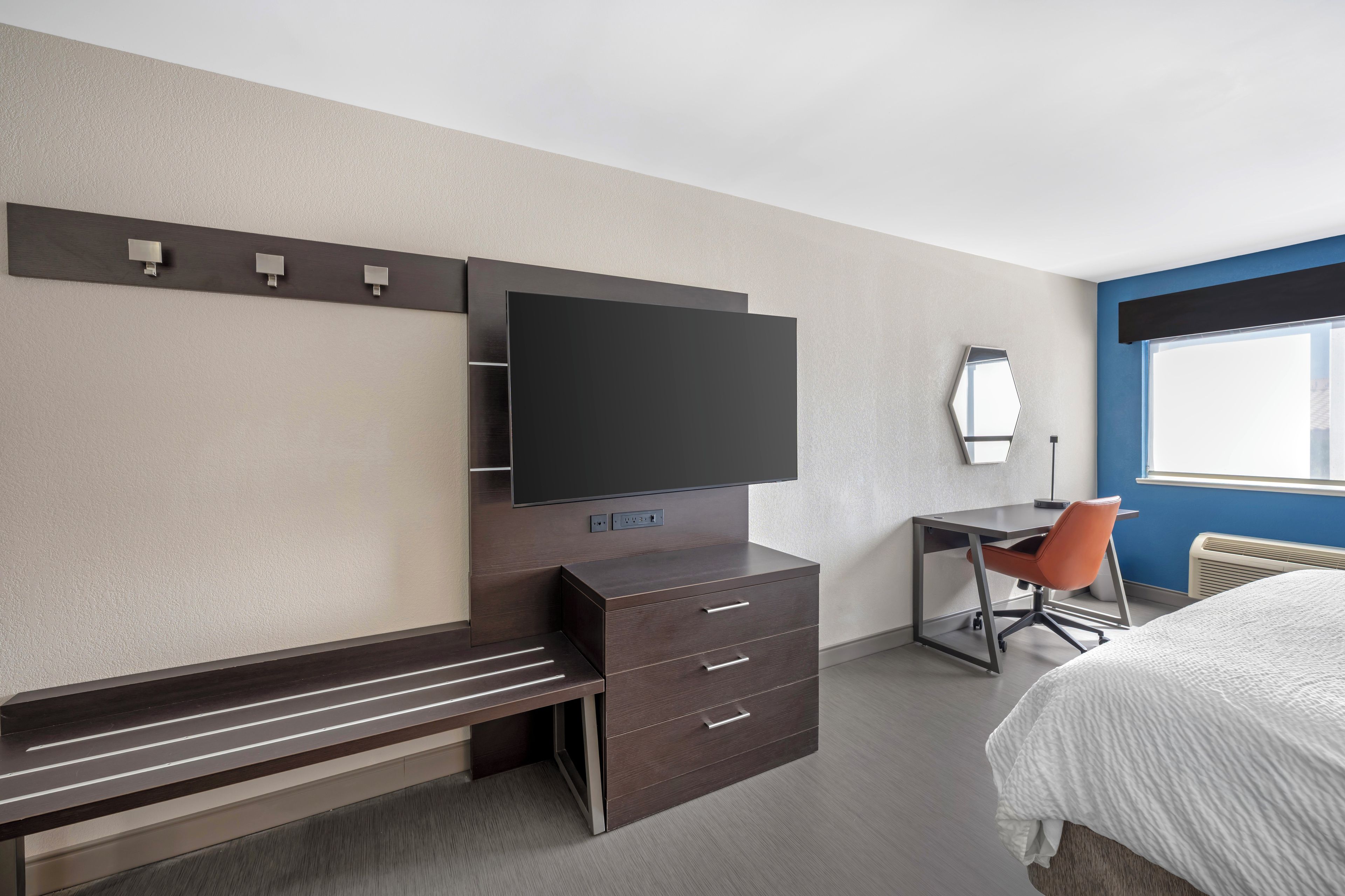 A hotel room with a bed, a desk, a chair, a TV mounted on the wall, and a window.