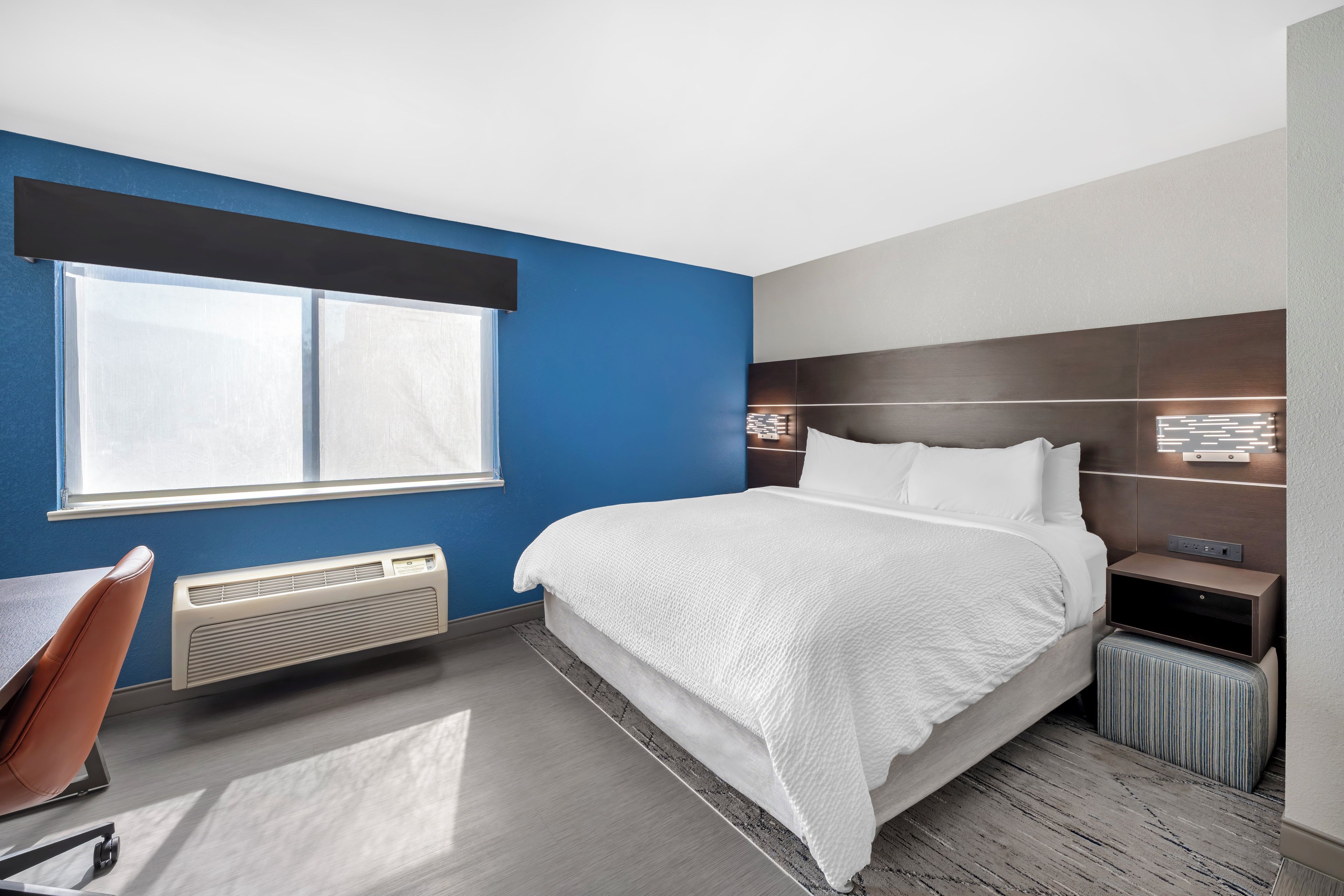 A hotel room with a large bed, a desk, a chair, a window, and a wall-mounted heater.