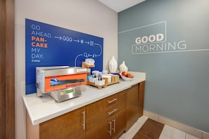A breakfast area with a flat-screen TV displaying a breakfast menu, a coffee machine, and a bowl of fruit on the counter. - Holiday Inn Express - Balloon Fiesta Park by IHG (Albuquerque)