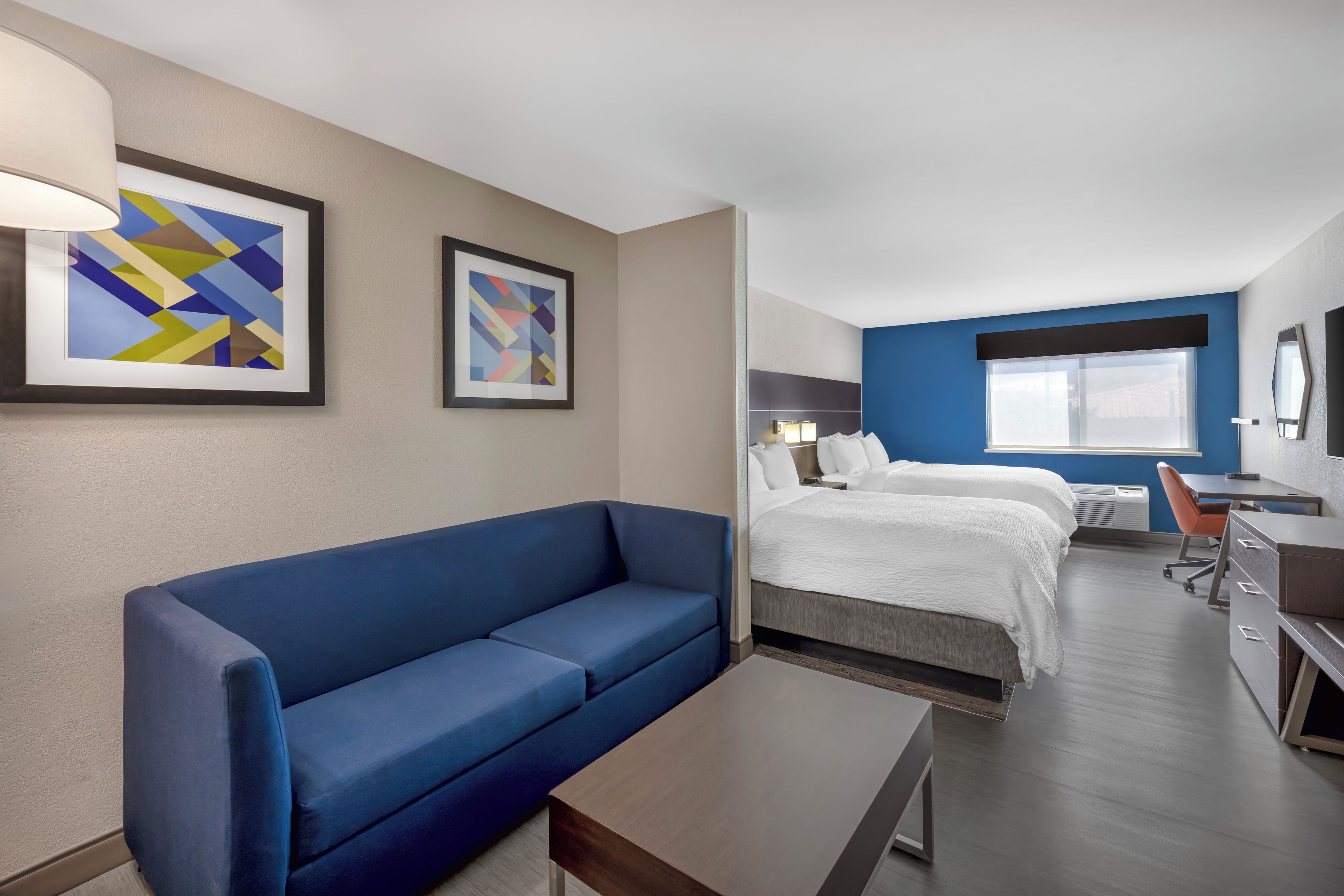 Een hotelkamer met een blauwe bank, een bed met wit bedlinnen, een bureau met stoel en twee abstracte schilderijen aan de muur.