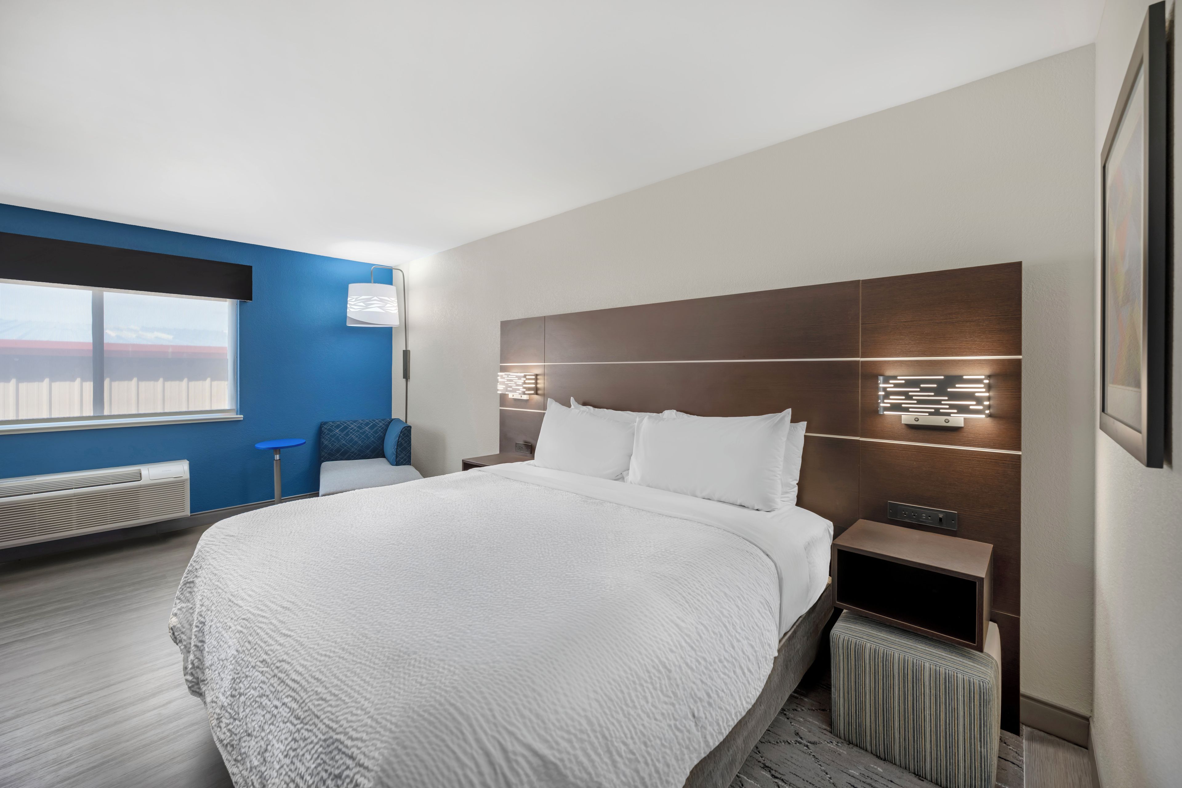 Ein modernes Hotelzimmer mit einem großen Bett, einem Nachttisch, einer Wandleuchte und einem Fenster mit Jalousien.