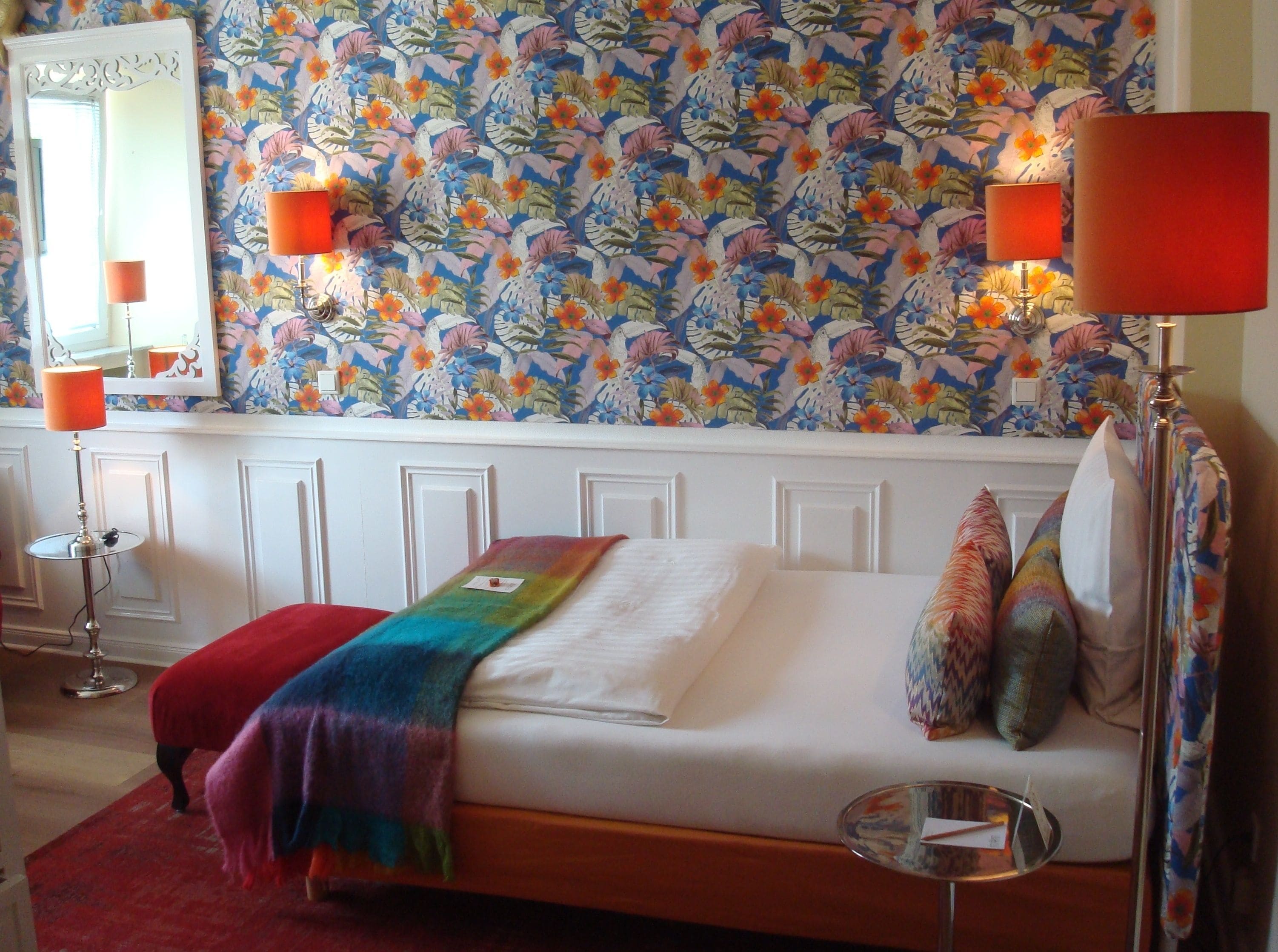 A bedroom with a floral wallpaper, a bed with a colorful blanket, two bedside lamps, and a mirror above the bed.