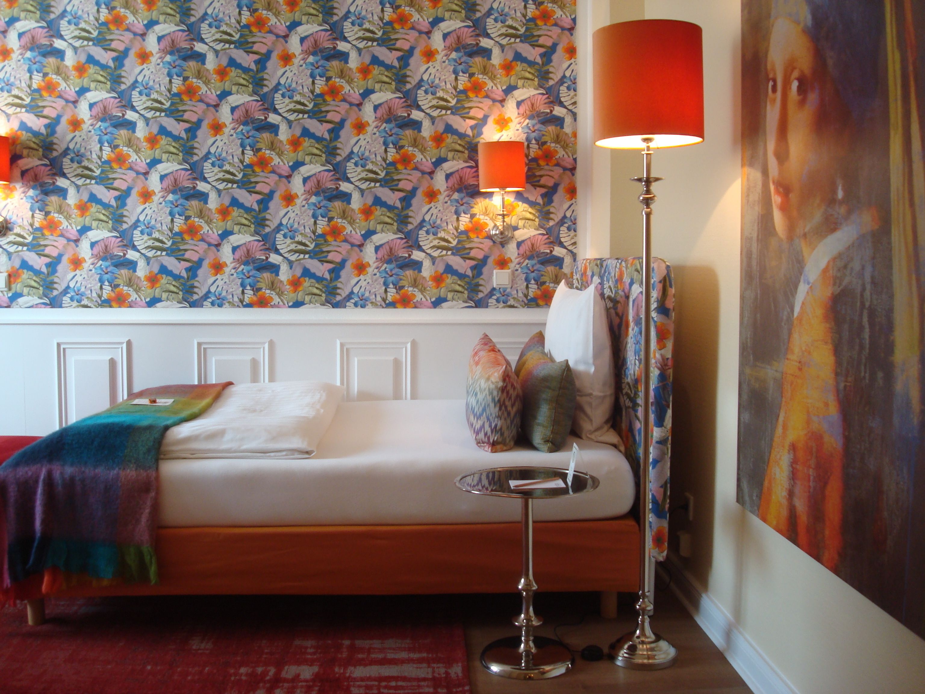 Un dormitorio con papel tapiz floral, una cama con manta colorida, una mesita de noche con lámpara y un cuadro en la pared.