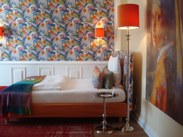 A bedroom with a floral wallpaper, a bed with a colorful blanket, a bedside table with a lamp, and a painting on the wall. - Hotel Sir & Lady Astor (Düsseldorf)
