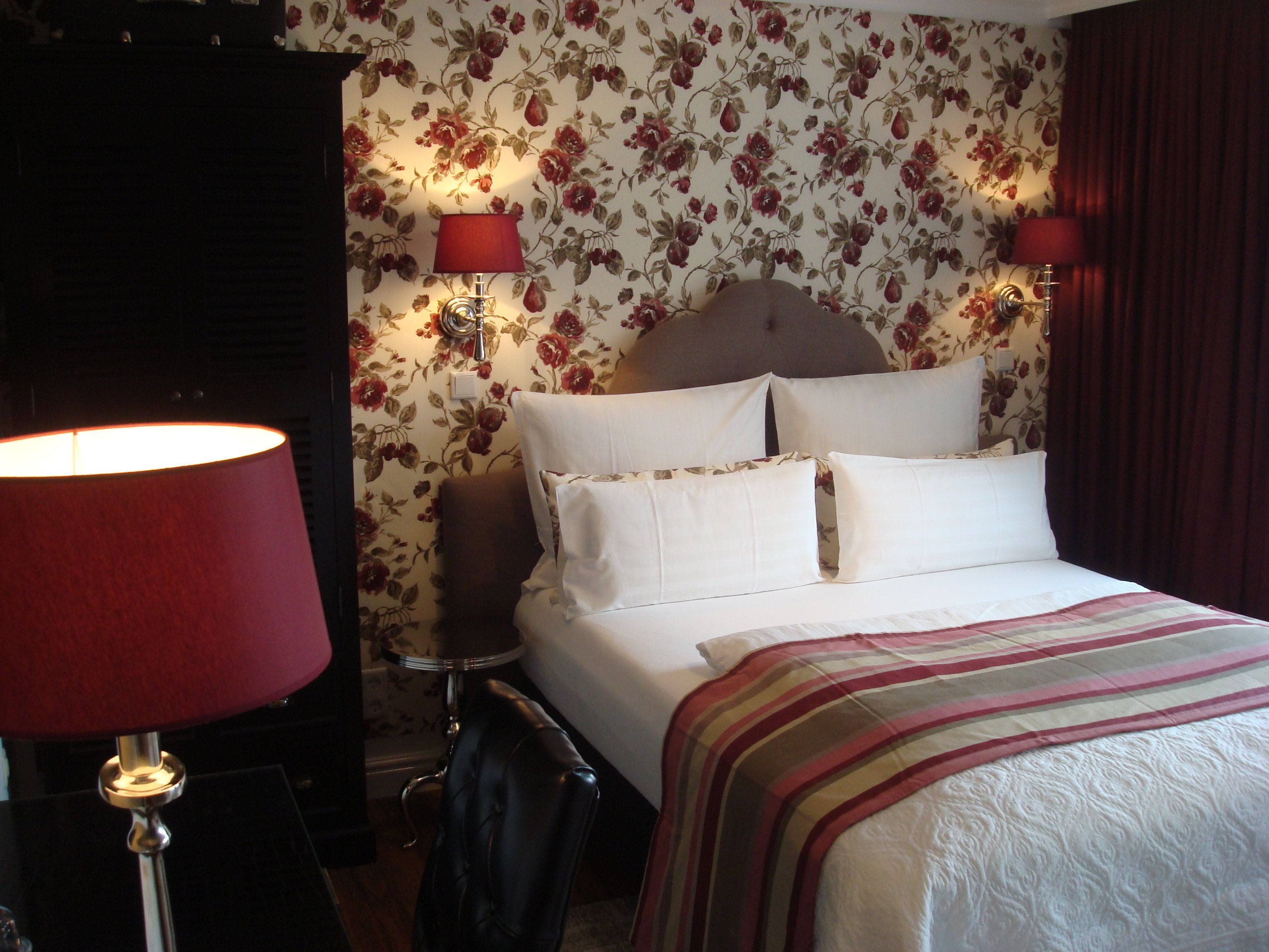 Ein Hotelzimmer mit einem Bett, zwei Nachttischlampen, einem Sessel und blumigem Wandpapier.