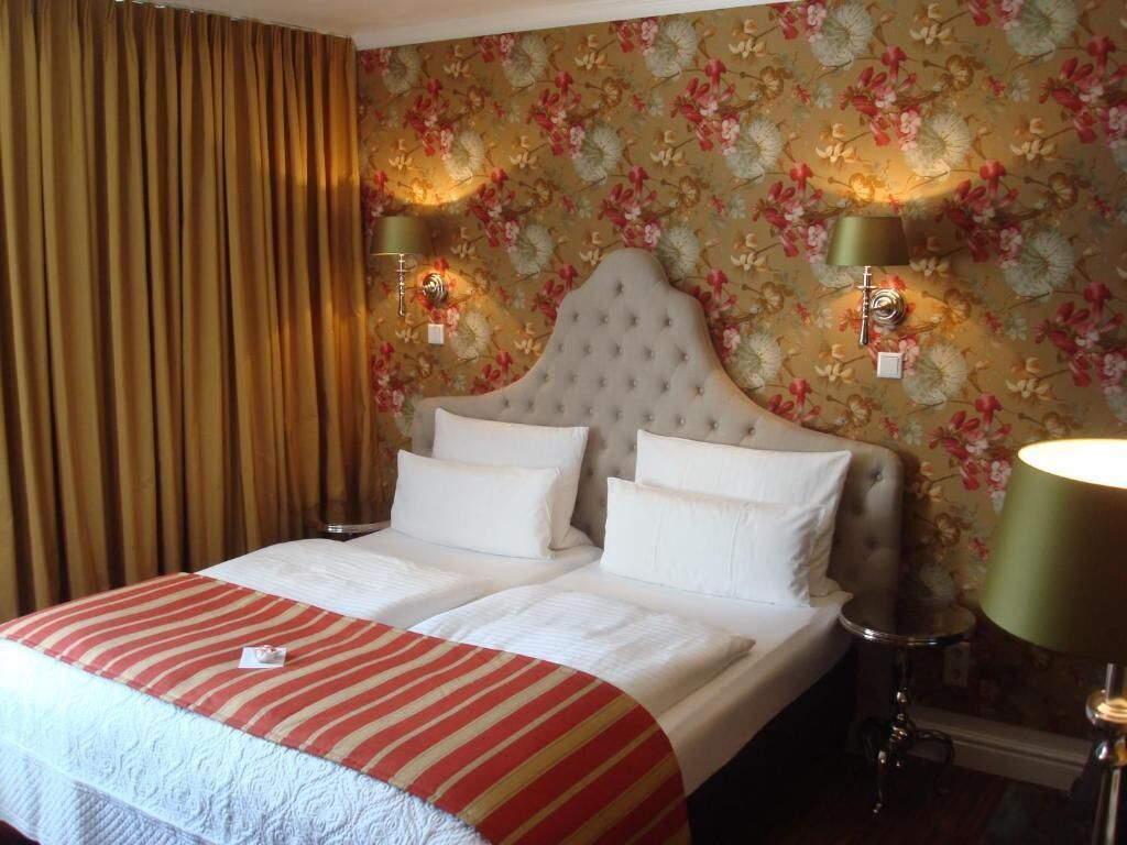 A hotel room with a large bed, two bedside lamps, floral wallpaper, and a curtain.
