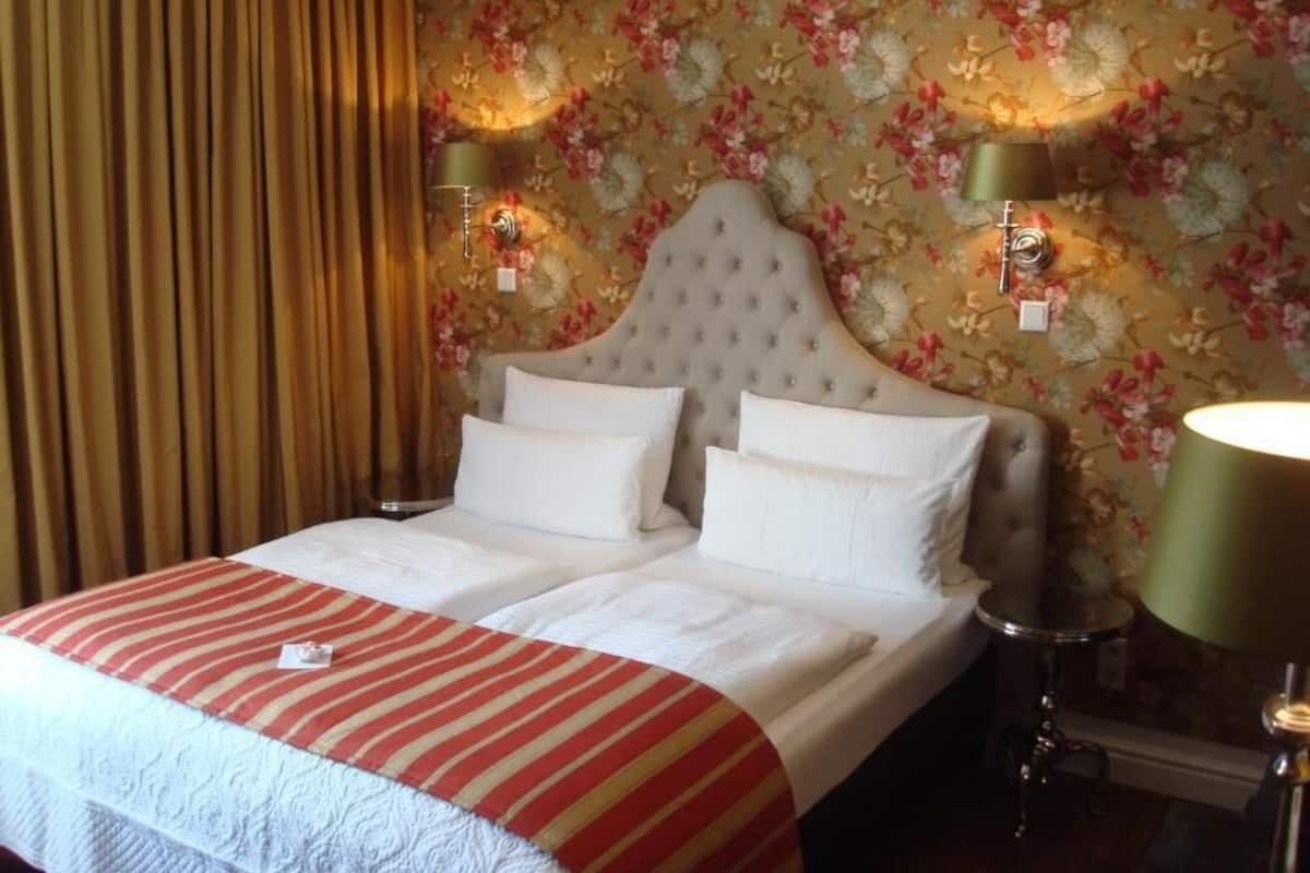 A hotel room with a large bed, two bedside lamps, floral wallpaper, and a curtain.