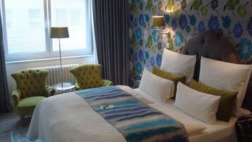 A hotel room with a bed, a green armchair, a small round table, a lamp, and a radiator.
