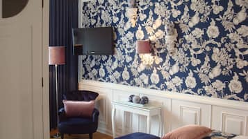 A bedroom with a floral wallpaper, a television mounted on the wall, a small table with decorative items, a chair, and a bed with a floral bedspread.
