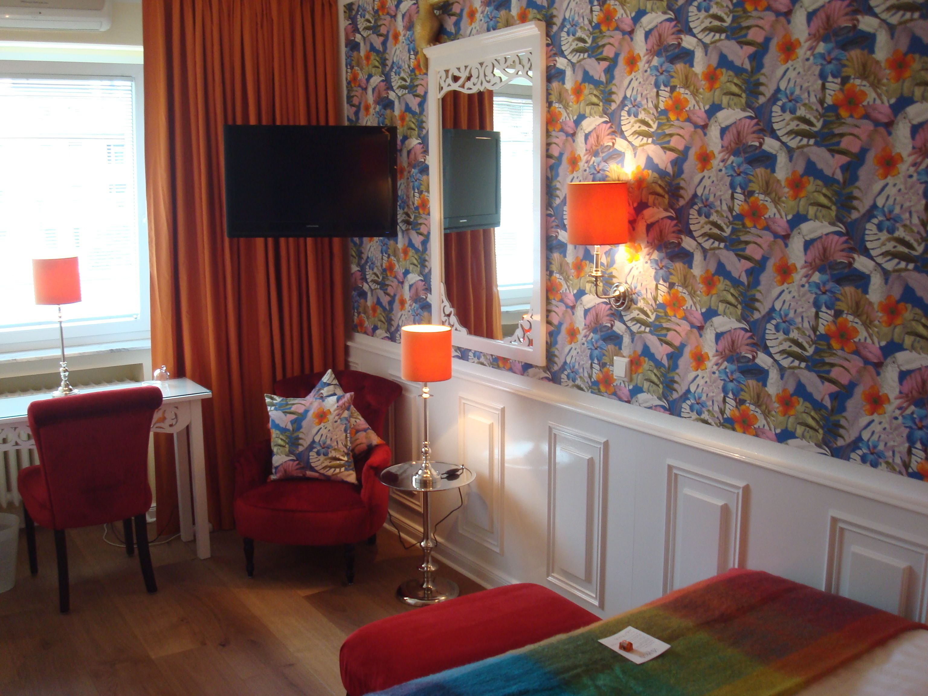 A hotel room with a red chair, a desk with a lamp, a television, and a colorful floral wallpaper.