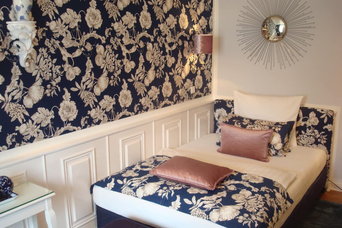 A bedroom with a floral wallpaper, a round mirror, and a bed with multiple pillows.