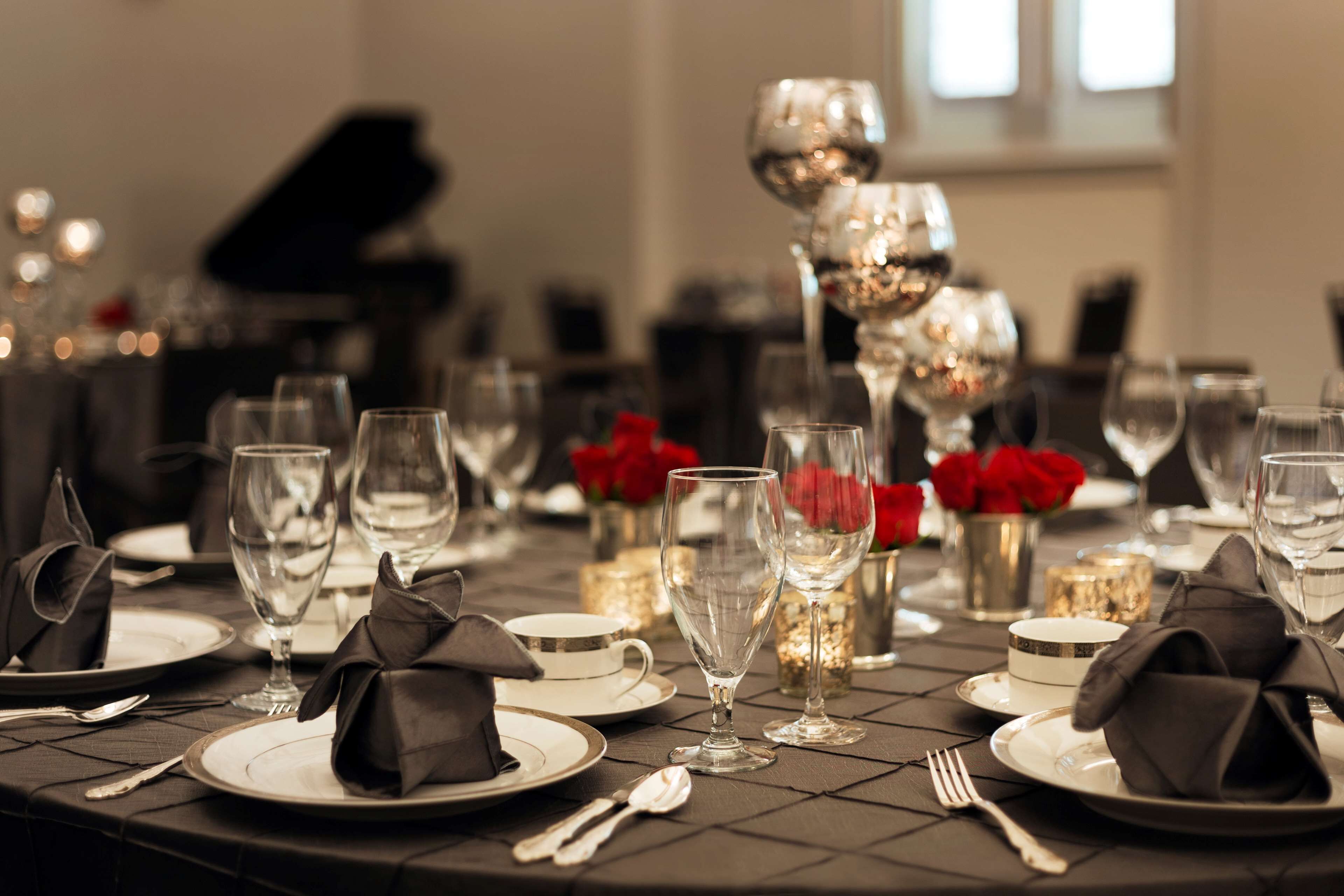 A formal dining table set with white plates, silverware, and clear wine glasses, accompanied by black napkins and red roses.