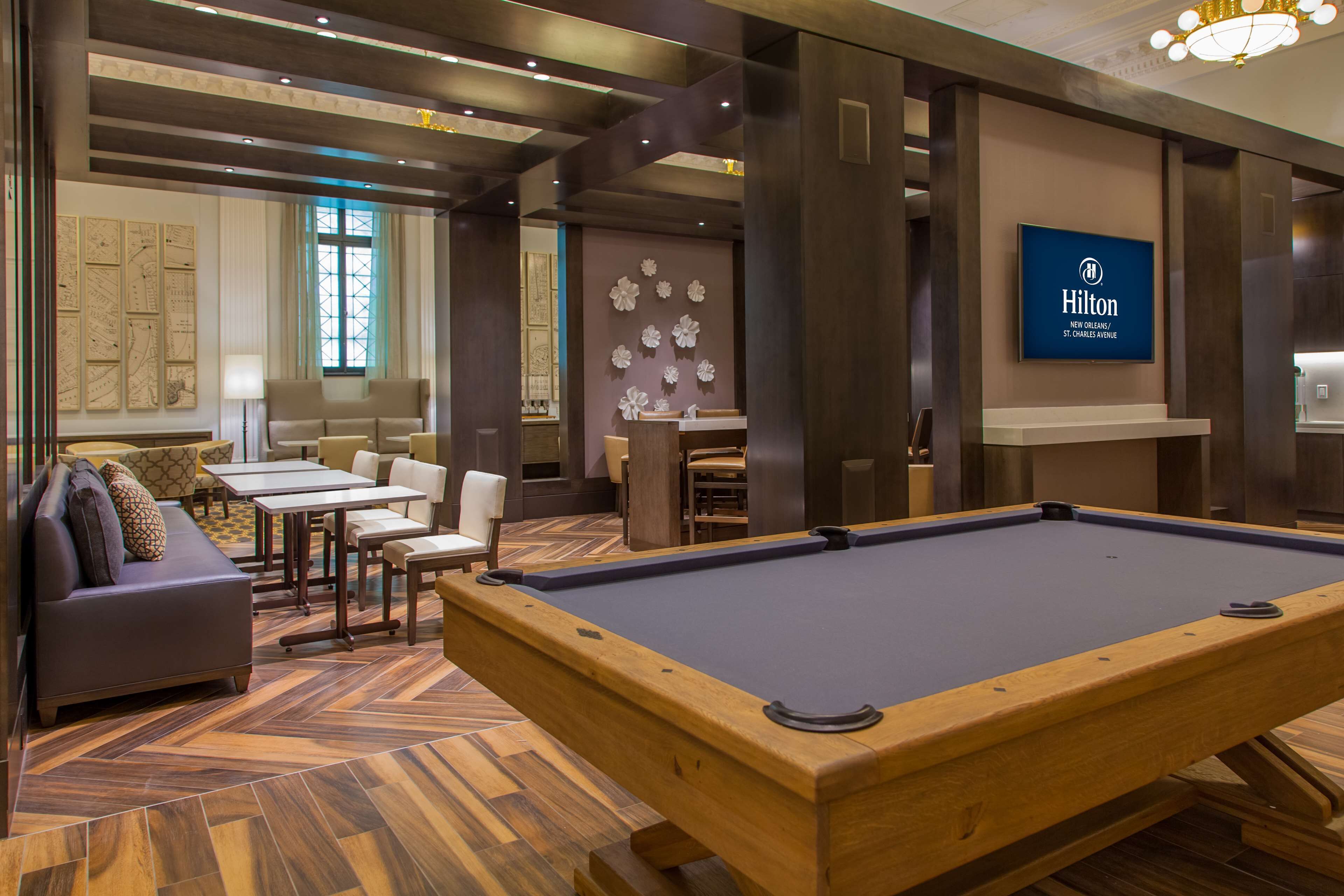 A pool table in a hotel lobby with a seating area, a large wall clock, and a sign for Hilton St. Charles Avenue.