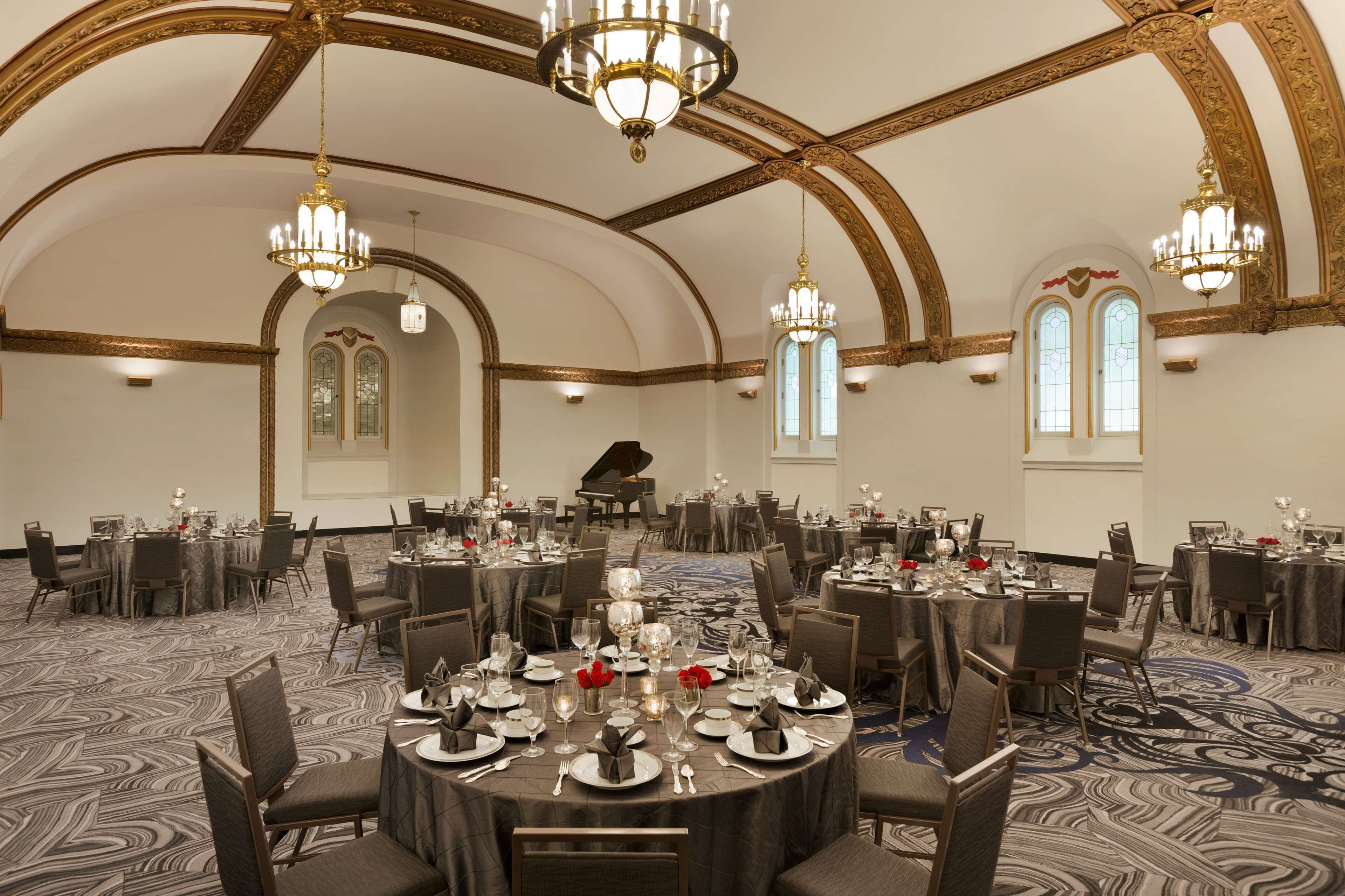 Une salle de banquet avec des tables rondes dressées pour un événement formel, agrémentée de lustres et d’un grand piano en arrière-plan.
