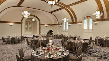 Une salle de banquet avec des tables rondes dressées pour un événement formel, agrémentée de lustres et d’un grand piano en arrière-plan.