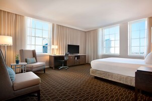 Habitación de hotel con cama, escritorio con televisor, silla y una pequeña mesa con lámpara.