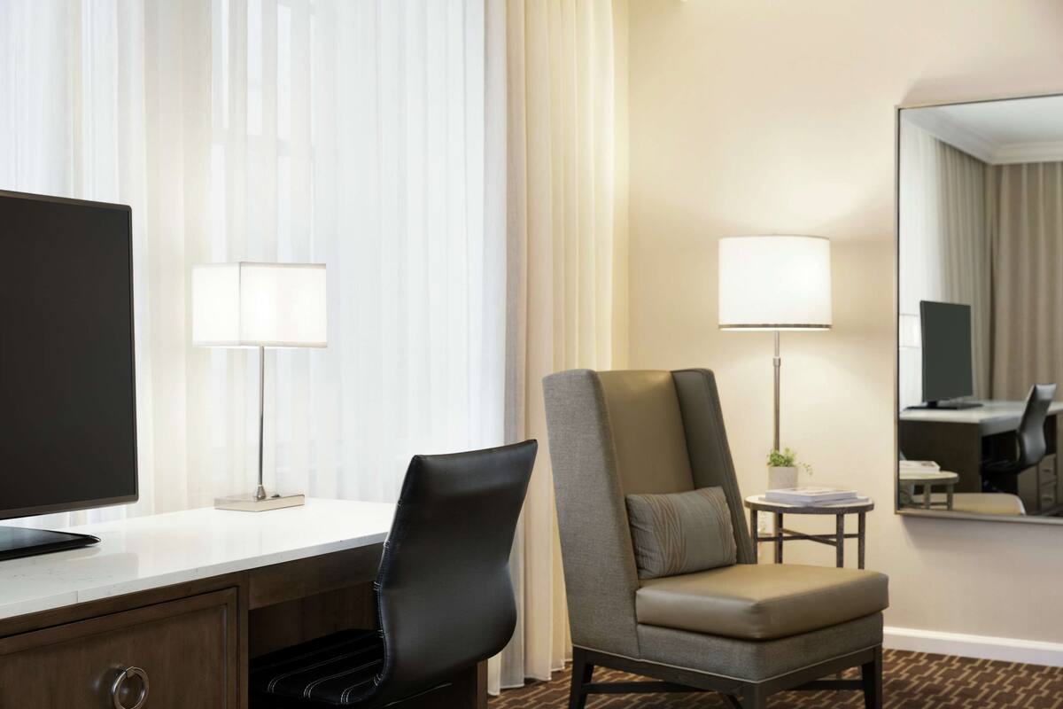 Habitación de hotel con televisor de pantalla plana, un escritorio con lámpara, un sillón cómodo, una mesita auxiliar con lámpara y un espejo en la pared.