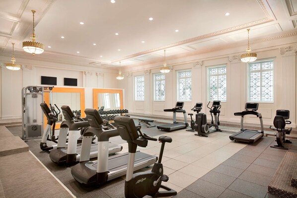 Un gimnasio amplio con diversos equipos de ejercicio, ventanas grandes y elegantes candelabros.