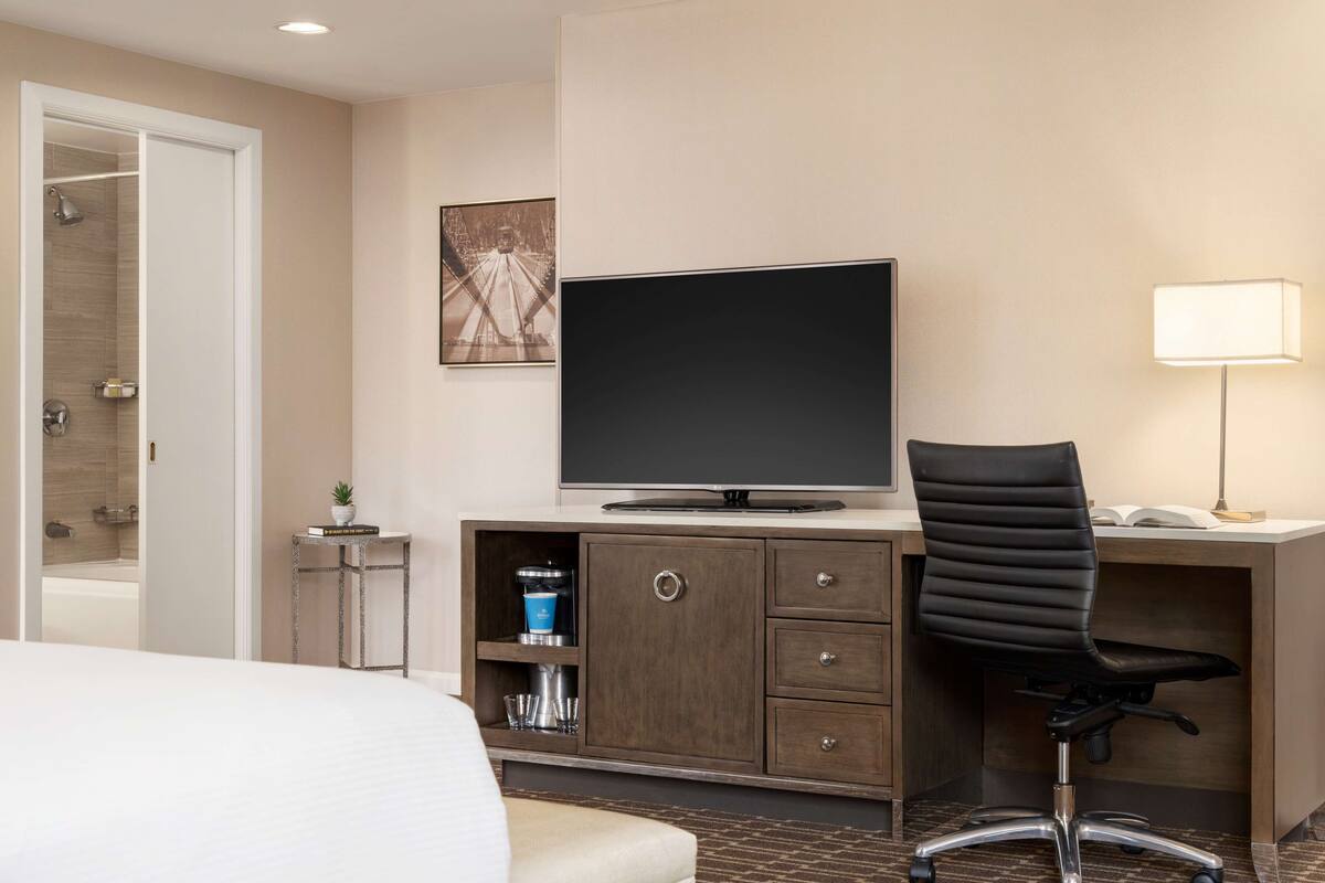 Habitación de hotel con una cama, un escritorio con computadora, una silla, un televisor y un mueble pequeño con artículos de tocador.