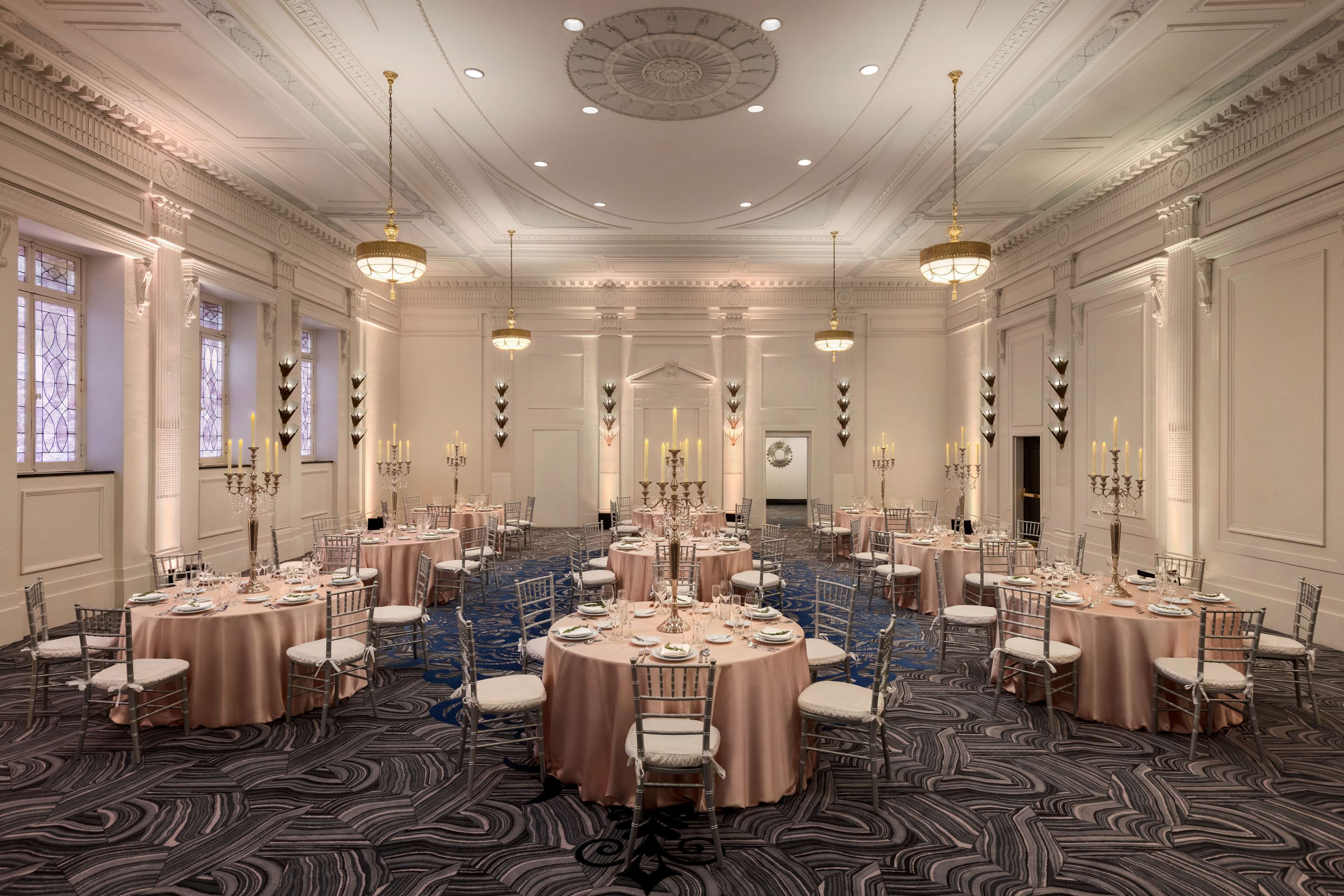 Un vaste hall de banquet avec des tables rondes, des lustres et des détails décoratifs élaborés au plafond.