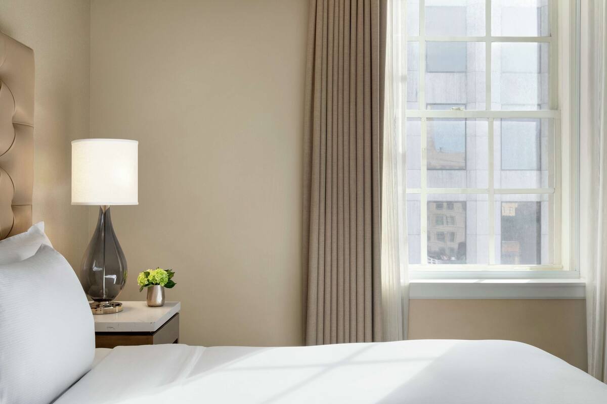 Habitación de hotel con cama, mesita de noche con lámpara, jarra con flores y ventana con cortinas.