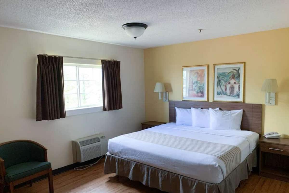 A hotel room with a large bed, a chair, a desk, and a window with curtains.
