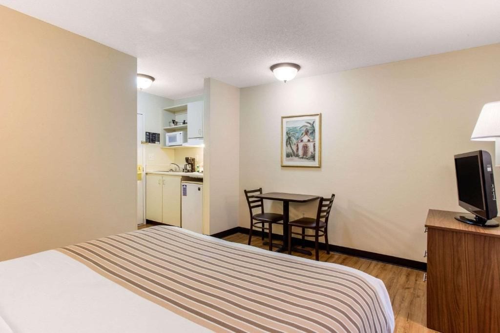 A hotel room with a bed, a small dining table with chairs, a television on a wooden stand, and a kitchenette in the background.