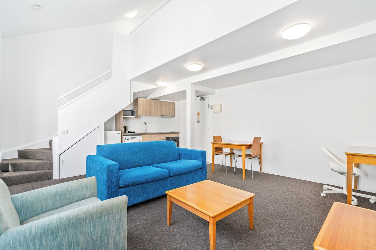 A modern living room with a blue sofa, a wooden coffee table, and a dining area with a table and chairs. A kitchen is visible in the background.