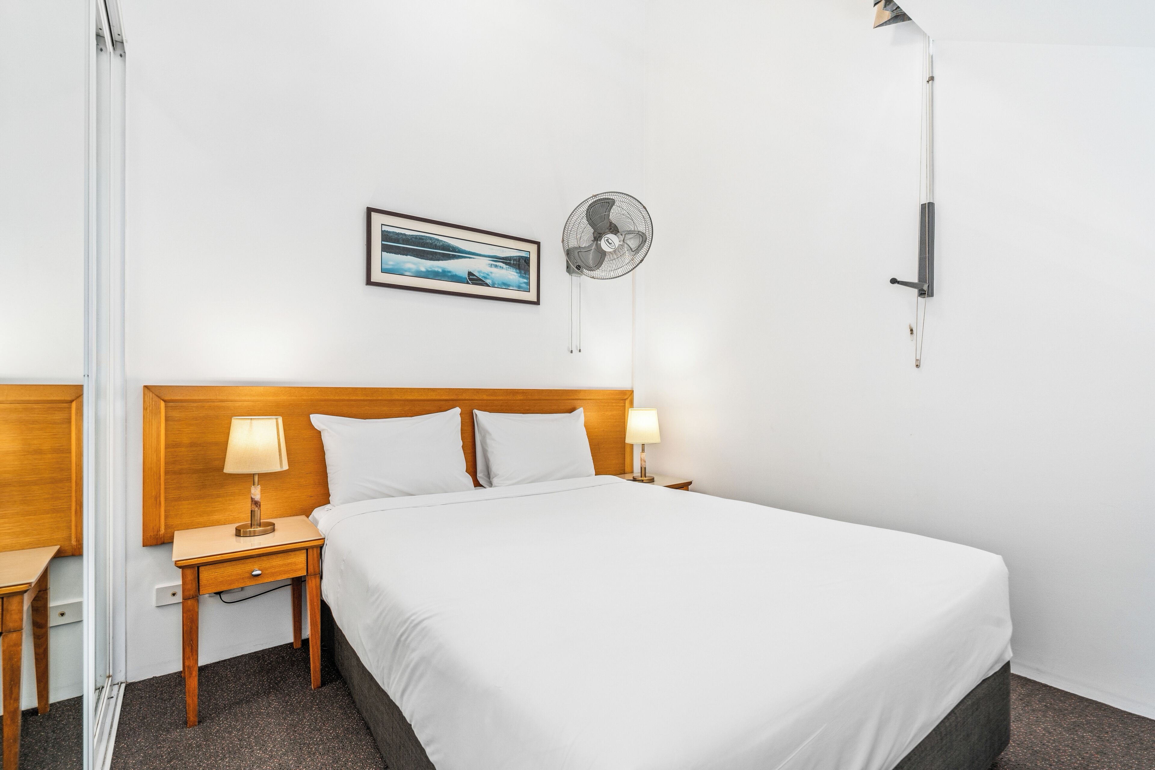 A hotel room with a large bed, wooden headboard, two bedside tables with lamps, a wall-mounted fan, and a framed picture on the wall.