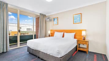 Habitació d'hotel amb un llit gran, una porta corredissa de vidre a un balcó, una petita taula de nit amb una làmpada i dos quadres enmarcats a la paret.