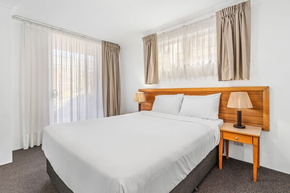 A hotel room with a large bed, a wooden nightstand, a lamp, and sheer curtains.
